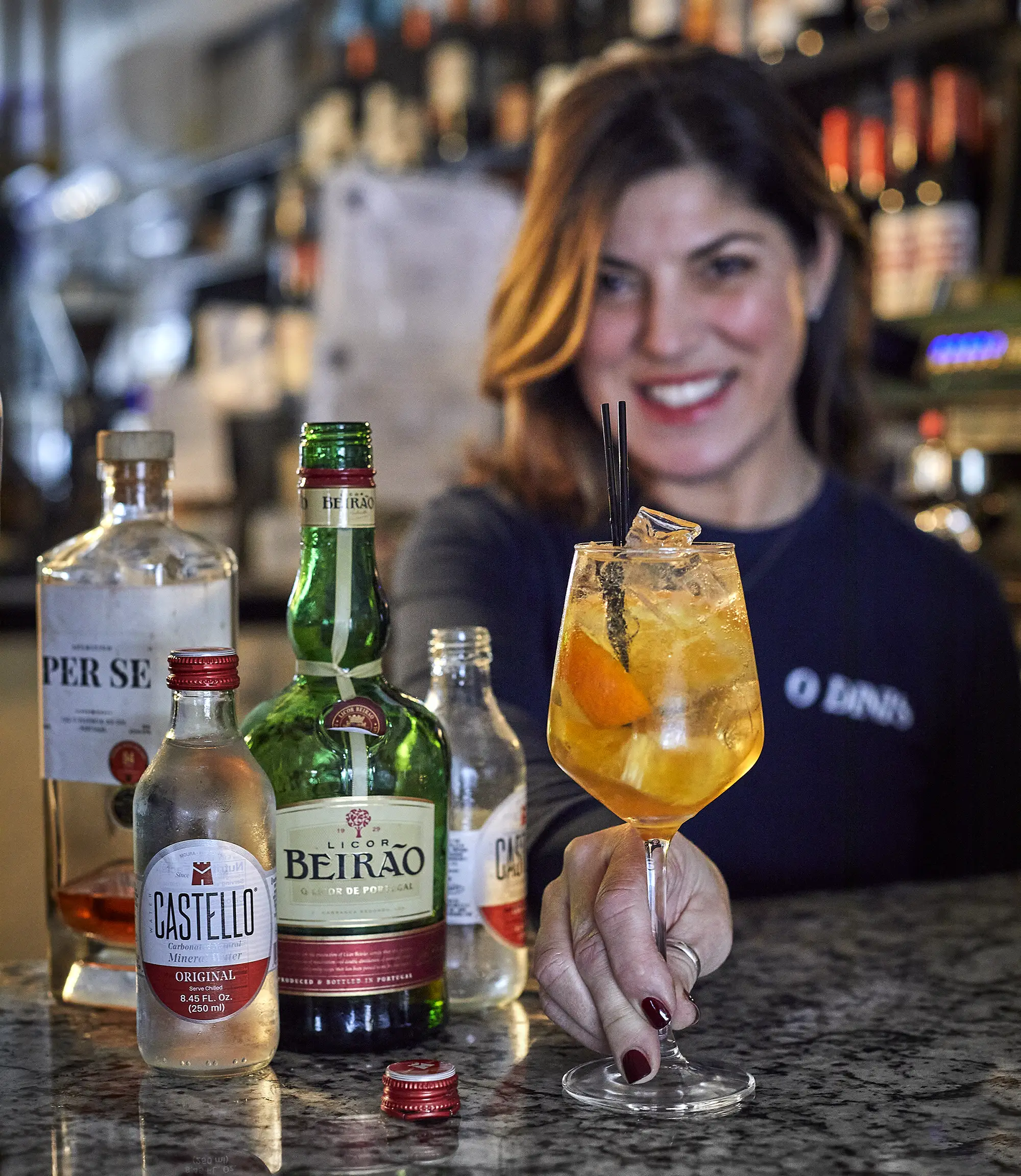 Woman presents orange mixed drink next to bottles of Portuguese alcohol